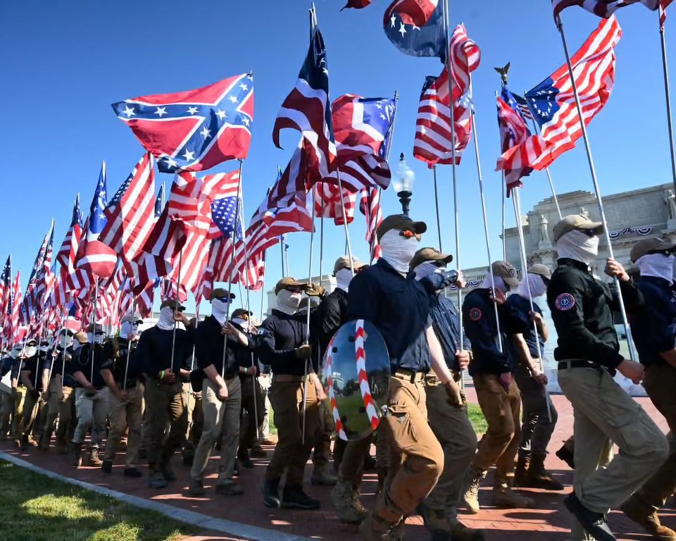 A group of Patriot  Front's people wearing similar clothing and masks are marching while holding a mix of Confederate and American flags.